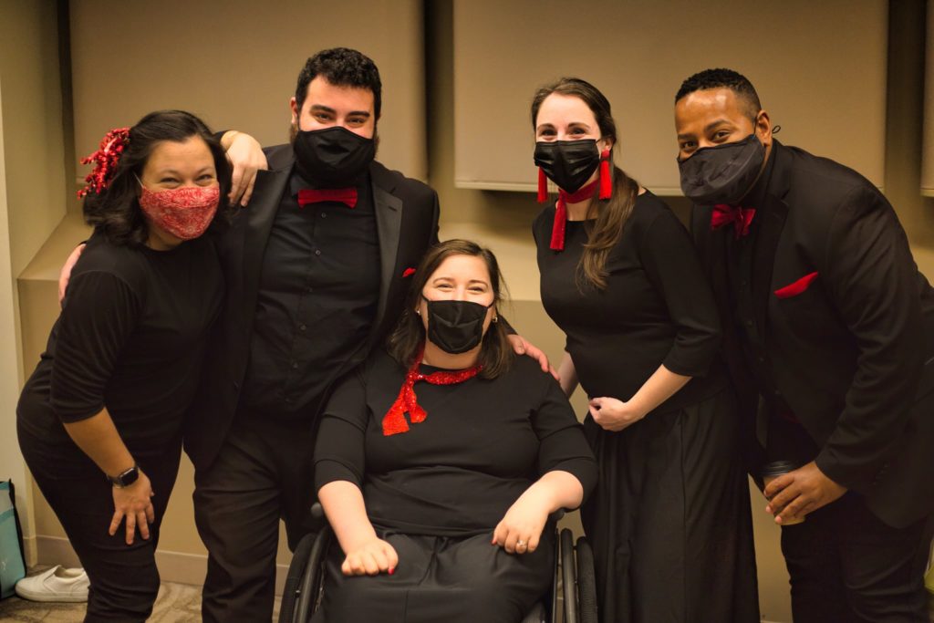 Five singers wearing black clothes and red accessories smile at you through their masks.